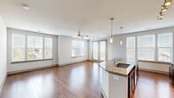 This single-floor residence offers a thoughtfully designed open-concept layout that seamlessly connects the kitchen, dining, and living areas. Rich hardwood flooring flows throughout the main living spaces, complemented by neutral-toned walls that create a clean, modern aesthetic. The kitchen is a standout feature, showcasing deep espresso-toned cabinetry extending to the ceiling, a classic subway tile backsplash, stainless steel appliances including a refrigerator, range, and microwave, and an expansive granite-topped island with an undermount sink and elegant pendant lighting overhead. Track lighting further illuminates the kitchen workspace. The open living area is bright and spacious, benefiting from multiple large windows dressed with horizontal blinds and a set of French doors that open to an outdoor balcony, inviting natural light and exterior views. A ceiling fan provides additional comfort to this area. The bedrooms feature plush carpeting, ceiling fans, and generous windows overlooking greenery. The bathroom is finished with large-format tile flooring, a dual-sink granite vanity with rich wood cabinetry, and a walk-in glass-enclosed shower. Additional utility space with carpeting and an electrical panel is also present within the unit.