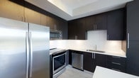 This single-floor space showcases a refined, contemporary design aesthetic centered around a well-appointed kitchen. The kitchen is a standout feature, finished with floor-to-ceiling dark espresso cabinetry that provides generous storage while creating a bold visual contrast against the crisp white walls and backsplash. The cabinetry is fitted with sleek brushed chrome hardware, adding a polished, modern touch throughout.

The kitchen is fully equipped with a suite of stainless steel appliances, including a French door refrigerator, an electric range with oven, an over-the-range microwave, and a built-in dishwasher — offering both functionality and a cohesive, professional look. A stainless steel undermount sink with a modern faucet is neatly integrated into the counter, complementing the overall design.

Light-toned flooring runs throughout, providing a bright, clean contrast to the dark upper and lower cabinetry. A recessed tray ceiling detail adds architectural depth and dimension to the kitchen area, enhancing the sense of height and openness.

The overall ambiance of this space is sleek and contemporary, with a carefully balanced palette of dark, white, and metallic tones that create a sophisticated and highly functional environment.