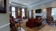 Located at 100 Saddle Cove Road in Bohemia, New York, this single-floor space exudes a refined, traditional elegance throughout its thoughtfully appointed interior. The main living area serves as the focal point of the space, showcasing a sophisticated design anchored by a stately white fireplace with an ornate marble surround. Rich crimson upholstered sofas and plaid accent chairs are arranged atop a decorative area rug, creating a warm and inviting seating environment. The room is framed by tall, floor-length draping curtains in warm earth tones, which complement the soft sage-green walls and classic wainscoting details. A tray ceiling with recessed lighting adds architectural depth overhead, while elegant wall sconces provide a soft, ambient glow. Large windows along the perimeter allow natural light to filter in, offering pleasant views of the surrounding greenery outside. A wall-mounted flat-screen television is positioned above the fireplace, blending modern convenience with the room's classic aesthetic. Stone tile flooring grounds the space with a durable yet stylish finish. The overall ambiance is one of comfort and understated luxury, making it a welcoming and functional environment well-suited for relaxed gatherings and everyday use.