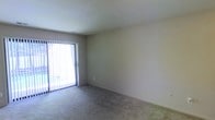 This single-floor residential space is designed with a straightforward and functional layout. The main living area is finished with wall-to-wall carpeting in a neutral tone, providing a comfortable and cohesive foundation throughout the space. The walls are painted in a soft, light beige, contributing to a clean and understated aesthetic.

A prominent feature of the living area is a full-width sliding glass door dressed with vertical blinds, which serves as both a source of natural light and a direct access point to an outdoor area. When the blinds are open, the door allows daylight to filter generously into the interior, brightening the room and creating a connection to the outside environment.

The ceiling is smooth and finished in a standard height, maintaining an open and uncluttered feel. A centrally placed ventilation register is visible, indicating a built-in HVAC system for climate control. Electrical outlets are positioned along the walls, offering practical utility throughout the space.

Overall, the space presents a clean, move-in-ready environment with a neutral palette that allows for flexible interior styling. The combination of soft carpeting, natural light access, and a simple, well-maintained finish makes this a functional and adaptable living space.