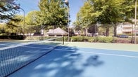 Located at 100 Preston Woods Trail in Sandy Springs, Georgia, this outdoor recreational facility features multiple tennis courts designed for year-round play. The courts are surfaced in a classic blue and green hard-court finish, clearly marked with crisp white lines and equipped with regulation nets. Each court is enclosed by sturdy chain-link fencing, providing both safety and containment during active play. Tall light poles are positioned throughout the facility, extending the availability of the courts into evening hours. The complex is framed by a natural canopy of mature trees, including towering pines and broadleaf hardwoods, creating a pleasant, shaded atmosphere that softens the surrounding environment. A brick retaining wall borders one section of the facility, adding a structured architectural element to the landscape. The courts are set within a well-established residential community, with neighboring buildings visible in the backdrop. The combination of ample greenery, solid infrastructure, and multiple playing surfaces makes this a well-appointed recreational amenity suited for both casual and competitive tennis.