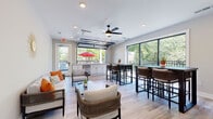 Located at 14735 Stable Gate Pl in Louisville, KY, this single-floor community or amenity space is thoughtfully designed for relaxed gathering and socializing. The interior is anchored by light-toned hardwood flooring that runs throughout, complementing the clean, neutral wall palette and white trim that give the space a fresh, contemporary feel.

A defining architectural feature is the glass-panel roll-up garage door along one wall, which opens seamlessly to an outdoor balcony area, blurring the line between indoors and out. Floor-to-ceiling sliding glass windows along an adjacent wall flood the space with abundant natural light and frame lush, tree-lined outdoor views. Recessed ceiling lighting and a centrally mounted ceiling fan with integrated light fixture ensure comfort and visibility in all conditions.

The layout is divided into two functional zones. A lounge area features woven rattan seating with cushioned sofas and chairs arranged around a low-profile coffee table, creating an inviting space for casual conversation. Adjacent to this is a bar-height dining and gathering zone with two dark-framed high-top tables and coordinating wicker bar stools. A wall-mounted flat-screen television is positioned for easy viewing from both areas. A separate entry door provides additional access to the outdoor balcony, enhancing the space's connectivity with the surrounding environment.