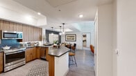 This single-floor residence showcases a thoughtfully designed open-concept layout that connects the kitchen, dining, and living areas into one cohesive, airy space. The kitchen is a standout feature, equipped with warm wood cabinetry, stainless steel appliances, a dark quartz countertop island with bar seating, and globe pendant lighting that adds a modern touch. A blue tile backsplash provides a subtle accent, while an in-unit washer and dryer add everyday convenience. The dining area sits adjacent, defined by a charcoal accent wall and overhead pendant lighting, flowing naturally into the living room. The living space features a ceiling fan, light wood-style flooring, and a welcoming, relaxed atmosphere. Two well-sized bedrooms each feature large windows that invite natural light and exterior views, with accent walls in muted tones adding depth and character. The primary bathroom includes a glass-enclosed shower and a floating wood vanity with dark countertop, while a secondary bathroom offers a tub-shower combination with clean white tile surround. Throughout, recessed lighting, neutral walls, and cohesive wood-toned finishes create a polished, contemporary ambiance.