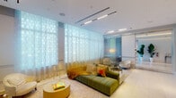 The lobby at 1818 Hollywood Blvd presents a refined, contemporary design that establishes an immediate sense of elegance upon entry. Automatic glass doors open to a bright, welcoming space where polished marble tile flooring—laid in both cream and contrasting darker tones—sets a clean, upscale foundation throughout. The entry corridor transitions seamlessly into a spacious central reception area distinguished by a striking circular coved ceiling with integrated ambient lighting, creating a warm, luminous focal point overhead.

A generously sized lounge area anchors one side of the lobby, featuring floor-to-ceiling patterned sheer drapery that diffuses abundant natural light while offering views of the surrounding landscaping. The seating arrangement includes a tufted emerald green sofa, cream sectional, and accent chairs with colorful throw pillows, composing an inviting, design-forward atmosphere. Additional seating vignettes with armchairs and side tables are thoughtfully positioned near the glazed exterior walls.

Tall tropical foliage in white planters adds organic warmth throughout the corridors, while panel-detailed walls in muted gray tones contribute architectural texture. Elevator access and well-lit passageways connect to further interior areas, completing a polished, cohesive lobby experience.