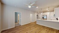 This single-story space presents a clean, contemporary design characterized by neutral tones, crisp white trim, and a cohesive finish throughout. The open-concept living and kitchen area serves as the heart of the unit, featuring warm wood-look luxury vinyl plank flooring, recessed lighting, and a ceiling fan that enhances the sense of airiness. A peninsula island anchors the kitchen, offering additional counter space and a built-in sink, while white shaker-style cabinetry extends to the ceiling for ample storage. Stainless steel appliances—including a refrigerator, range, microwave, and dishwasher—complement the bright white countertops and under-cabinet lighting. Pendant lights hang above the island, adding a polished touch. The living area connects seamlessly to the bedroom wing via interior doors, transitioning from hard flooring to plush carpet. The bedroom is well-proportioned with a ceiling fan and a window fitted with blinds for natural light control. The primary bathroom features large-format tile flooring, a wide vanity with a backlit mirror, and direct access to a generously sized walk-in closet equipped with dual hanging rods and shelving. The overall ambiance is modern, clean, and move-in ready.