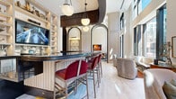 Located at 770 Juniper St NE in Atlanta's Midtown neighborhood, this sophisticated single-floor hospitality space combines classical architectural detail with contemporary design to create a refined and welcoming environment. The grand reception area anchors the space with soaring ceilings, light wide-plank hardwood flooring, and a striking circular mirrored ceiling installation overhead. A richly appointed front desk, accented in deep teal and dark wood tones, serves as the focal point of arrival. Adjacent to the entry, a dramatic floor-to-ceiling built-in shelving unit finished in warm red tones displays an eclectic collection of art and decorative objects, illuminated by a cascading crystal chandelier. The lounge area features elegant panel-molded walls, plush seating arrangements, and a large statement fireplace, creating an intimate and inviting atmosphere. A curved bar with a dark stone countertop, velvet crimson bar stools, and globe pendant lighting offers a stylish social hub, complete with arched architectural detailing and integrated display shelving. Comfortable dining nooks with banquette seating and curated artwork provide versatile gathering spaces. A fully equipped back-of-house kitchen with professional-grade stainless steel appliances supports event and hospitality services. Natural light streams in through tall windows, enhancing the overall sense of openness and sophistication throughout.