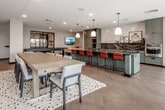 Located on Broadway Boulevard in Kansas City's Midtown neighborhood, this expansive single-floor amenity space is thoughtfully designed to serve as a social and entertainment hub. Herringbone-patterned hardwood flooring flows throughout, anchoring a cohesive aesthetic that blends modern and transitional design elements.

The fully equipped demonstration kitchen serves as the heart of the space, featuring sage-green cabinetry, dark stone countertops, a butcher block island, built-in appliances, and pendant lighting overhead. A long bar counter lined with rust-orange upholstered stools provides ample seating for gathering and socializing.

Adjacent to the kitchen, a generous dining area accommodates large groups at a communal wood table, while café-style seating near floor-to-ceiling windows invites casual conversation with panoramic views of Broadway Boulevard below. Glass doors connect the interior to an outdoor pool area, flooding the space with natural light.

The lounge area features a dramatic marble-surround gas fireplace flanked by artwork and a circular pendant chandelier, creating a warm, inviting atmosphere. Comfortable seating arrangements include a mix of sofas, armchairs, and a woven ottoman coffee table. A dedicated entertainment nook with a wall-mounted television and gallery wall adds another layer of function, while a billiards table rounds out the recreation offerings.