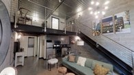 This two-story industrial loft makes a striking first impression with its raw, urban aesthetic and open-concept design. The main level showcases exposed concrete block walls, polished concrete floors, and a standout corrugated metal ceiling that adds an unmistakable industrial character to the space. Soaring double-height ceilings amplify the sense of volume and openness throughout.

The living area is anchored by a comfortable teal sofa and warm accent furnishings, creating an inviting contrast against the space's rugged architectural backdrop. A statement sputnik-style chandelier adds a touch of contemporary flair overhead. The kitchen is seamlessly integrated into the main level, featuring grey cabinetry, stainless steel appliances, and a functional layout that keeps everything within easy reach.

A bold steel staircase with sleek cable railings leads to the mezzanine level above, which overlooks the main living space through an open railing system — ideal for use as a sleeping area or private retreat. The interplay between the raw industrial materials and carefully chosen furnishings gives this loft a distinctive, modern edge that balances functionality with style.