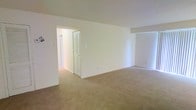 This single-floor residential space offers a straightforward and functional layout designed for comfortable everyday living. The main living area is generously sized, featuring wall-to-wall beige carpeting that adds warmth and softness underfoot. The walls are finished in crisp white, lending a clean, neutral backdrop that allows for versatile interior styling and decorating options.

The space benefits from an open feel, with smooth ceilings and simple, unobtrusive architectural lines that maximize the sense of roominess. A doorway connects the main living area to an adjacent hallway or secondary room, creating a natural flow throughout the floor plan. A classic bi-fold louvered closet door is visible along the hallway, providing practical storage access while contributing a traditional residential character.

Electrical outlets are thoughtfully placed along the baseboards, and standard light switches are positioned conveniently near doorways. The overall ambiance is calm and understated, reflecting a move-in ready condition with well-maintained surfaces. The neutral color palette and carpeted flooring make this space highly adaptable, offering a blank canvas suited to a variety of furnishing styles and personal preferences. The single-floor layout ensures ease of accessibility and navigation throughout the entire space.
