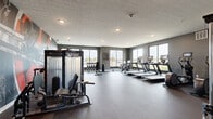 Located at 2101 Railway Ln. in Marysville, Ohio, this fitness center offers a well-appointed, single-floor exercise facility designed to support a comprehensive workout experience. The space is characterized by its modern aesthetic, with dark gray walls, durable rubber and concrete-style flooring, and recessed lighting throughout that creates a clean, focused atmosphere.

Large floor-to-ceiling windows line multiple walls, flooding the space with abundant natural light and providing open views of the surrounding exterior — an energizing backdrop for any workout. A striking, large-scale industrial mural serves as a bold visual centerpiece, adding a dynamic and motivating character to the room.

The fitness floor is thoughtfully organized into distinct functional zones. A dedicated cardio area houses elliptical machines, treadmills, a stair climber, and stationary bikes, each equipped with individual screens for entertainment. A strength training section features a cable machine system, weight benches, and a full rack of dumbbells ranging across various weights. Full-length mirrors along one wall enhance the sense of space and allow for proper form checks during exercise.

Glass partitions divide sections of the facility, maintaining visual openness while defining separate workout areas. The overall layout provides ample open floor space for movement, making this fitness center a well-rounded and inviting environment for residents and users of all fitness levels.