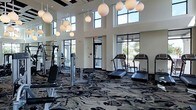 This single-story fitness center is designed with a contemporary aesthetic and a spacious, open layout that accommodates a variety of exercise equipment. The room is anchored by soaring double-height ceilings adorned with a series of oversized globe pendant lights, creating a stylish yet functional atmosphere. Large, floor-to-ceiling windows and transom windows line the perimeter walls, flooding the interior with abundant natural light and providing views of the surrounding outdoor environment. The boldly patterned carpet in dark tones of black, grey, and white adds visual interest while defining the workout area. The facility is equipped with cardio machines, including multiple treadmills, as well as strength-training equipment and free weights, making it suitable for a full-body workout. Full-length mirrors are strategically positioned along one wall to maximize the sense of space and assist users during exercise. The clean, neutral wall tones complement the modern finishes throughout, while the generous footprint ensures ample room between equipment for comfortable, unrestricted movement.