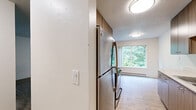 Located at 1820 NE 104th Ave in Portland, Oregon, this two-bedroom, one-bathroom unit offers a practical and comfortable single-floor layout. The galley-style kitchen is well-appointed with modern wood-toned cabinetry extending to the ceiling, light quartz-style countertops, stainless steel appliances including a range and refrigerator, and wood-look tile flooring. A window at the end of the kitchen frames views of the surrounding greenery, allowing natural light to brighten the space. The living area features warm wood-look flooring and a clean, open feel with neutral white walls. Both bedrooms are carpeted and finished with crisp white walls and sliding double-panel closet doors that maximize storage. Each bedroom includes a window with outdoor views and a baseboard heating unit. The full bathroom is bright and clean, featuring a combination tub and shower, wood-look tile flooring, a dark-toned vanity with a white countertop, and accessibility grab bars. A standout feature is the private wooden deck accessible from the unit, which overlooks a lush, tree-filled landscape — offering a serene outdoor retreat. The overall ambiance is clean, neutral, and move-in ready, with practical finishes throughout.