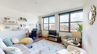 This well-designed single-floor urban residence offers a comfortable and functional layout bathed in natural light. The living area is a standout space, anchored by oversized windows with dark frames that frame expansive city views of the surrounding urban skyline. The open layout accommodates both a seating area and a dedicated workspace, with warm hardwood flooring running throughout and a soft area rug adding texture and warmth.

The kitchen is fully equipped and thoughtfully designed, featuring rich warm-toned wood cabinetry, dark granite countertops, a classic subway tile backsplash, and a full suite of stainless steel appliances including a refrigerator, range, microwave, and dishwasher. Light hardwood flooring and generous counter space make this kitchen as practical as it is attractive.

Two bedrooms offer comfortable retreats, each carpeted and featuring large windows that continue the city views seen throughout the unit. The rooms are well-proportioned with clean, neutral walls conducive to a variety of layouts.

The bathroom is clean and functional, finished with dark tile flooring, a combination tub and shower with sliding glass doors, a single vanity with an integrated sink, and a large mirror with overhead lighting. Every room in this residence benefits from abundant natural light and thoughtful urban views.