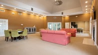 Located at 5700 Magnolia Chase Way in Virginia Beach, Virginia, this single-floor community clubhouse offers a well-appointed gathering space designed for comfort and versatility. The main lounge area is generously proportioned, featuring soaring vaulted ceilings with crown molding details and a coffered tray ceiling section that adds architectural depth. Warm, earth-toned walls complement the neutral carpeted flooring throughout, creating an inviting and cohesive atmosphere. Recessed lighting illuminates the space evenly, while multiple arched windows and sets of French doors flood the interior with natural light and provide views of the surrounding greenery. A classic white fireplace with a traditional mantel serves as a focal point on one wall, accompanied by a wall-mounted television, making it ideal for relaxed gatherings. A dining area with a dark-stained table and upholstered seating sits adjacent to the lounge, offering a defined space for group meals or meetings. A decorative sideboard with lattice-panel doors adds a refined touch. The kitchen features rich, dark espresso cabinetry, crisp white countertops, a double-basin sink, and stainless steel appliances including a full-sized refrigerator, providing full functionality for hosted events. French doors open directly to the outdoors, seamlessly connecting the interior to the exterior environment.