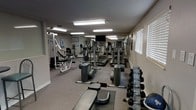 This fitness room is designed to support a well-rounded workout experience within a single-floor layout. The space is outfitted with multiple treadmills and elliptical machines positioned toward the center and back of the room, providing ample cardio options. A wall-mounted flat-screen television is centrally placed, visible from most cardio stations. Along the left side of the room, a multi-function cable and weight machine offers strength training versatility, accompanied by an adjustable bench for targeted exercises. A dumbbell rack lines the right side of the room, stocked with a range of free weights to accommodate various fitness levels. A flat workout bench sits in the foreground, suitable for stretching or floor exercises. Overhead fluorescent lighting ensures the space is consistently well-lit, complemented by natural light filtering through window blinds along the right wall. The flooring features a low-pile carpet, and the neutral-toned walls contribute to a clean, functional atmosphere. The layout is efficient, allowing users to move between equipment comfortably while maintaining a focused, no-frills fitness environment.