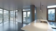 This single-floor space showcases a contemporary open-concept design with clean lines and industrial-modern influences throughout. The living and kitchen areas flow seamlessly together, anchored by a substantial kitchen island with a smooth white countertop and an integrated stainless steel sink fitted with a modern arc-neck faucet. Overhead, slender cone-shaped pendant lights descend from the ceiling, adding a sculptural focal point above the kitchen workspace.

A bold exposed concrete column serves as an architectural centerpiece, grounding the space while adding an industrial character that complements the polished dark flooring underfoot. The ceilings are high and uncluttered, contributing to an open, airy atmosphere throughout.

Natural light floods the interior through expansive floor-to-ceiling windows along the perimeter, framing sweeping urban city views. A set of French doors provides direct access to an outdoor balcony, further connecting the interior to the cityscape beyond. The overall ambiance is sophisticated and minimalist, blending raw structural elements with refined modern finishes to create a striking, cohesive living environment.