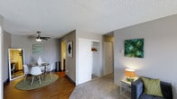 This single-floor unit offers a practical and well-organized layout designed to maximize every inch of available space. The living area features plush carpeting that transitions into hardwood-style flooring in the dining zone, creating a natural visual distinction between spaces. A ceiling fan overhead provides both function and a subtle design element, while neutral gray walls offer a versatile backdrop throughout.

An open coat closet with a hanging rod is conveniently positioned near the entryway, providing accessible storage without consuming additional square footage. Adjacent to the living space, a compact dining area accommodates a small round table with modern white chairs, set atop a circular area rug that anchors the space. A textured popcorn ceiling adds a classic residential character overhead.

Wall art featuring abstract green tones adds pops of color against the neutral palette. A partially visible hallway leads toward what appears to be a bathroom and additional rooms, suggesting a functional floor plan. The overall ambiance is clean, understated, and move-in ready, with a straightforward design suited to everyday living.