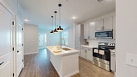 Located at 9292 Grand Cordera Pkwy in Colorado Springs, CO, this one-bedroom, one-bathroom unit offers a thoughtfully designed single-floor layout. The kitchen serves as the centerpiece of the living area, showcasing a large white waterfall-edge island with an undermount sink, sleek gray cabinetry extending to the ceiling, a herringbone tile backsplash, and a full suite of stainless steel appliances including a refrigerator, electric range, and over-the-range microwave. Three stylish matte black pendant lights illuminate the island, adding a modern touch. Warm wood-look flooring runs throughout the main living and kitchen areas, creating a cohesive and inviting atmosphere. Natural light filters in through the windows, brightening the open-concept space. The bathroom features a full tub/shower combination accented by large-format marble-look tile surround, paired with wood-look flooring. The bedroom is finished with plush carpet, a ceiling fan, and a generously sized window offering exterior views. A dedicated walk-in closet with built-in shelving and hanging rods provides ample storage. Clean lines, neutral tones, and modern finishes define the overall aesthetic of this well-appointed Colorado Springs residence.