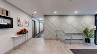 Located at 855 Jervis St in Vancouver's West End, this building lobby offers a well-appointed and functional entry experience for residents. The space is finished with large-format light stone tile flooring that extends throughout, creating a clean and cohesive aesthetic. A standout geometric feature wall clad in a soft sage-toned paneling with a diagonal diamond pattern serves as a striking architectural focal point near the staircase, which is equipped with stainless steel handrails. The entrance is framed by floor-to-ceiling glass doors that invite natural light and provide views to the streetscape outside. Recessed pot lighting illuminates the space evenly throughout. Residents are greeted by a modern Sworindi smart parcel locker system positioned prominently near the entrance, offering a convenient package retrieval solution. A dedicated mailbox alcove provides individual mail compartments in an organized niche just off the main lobby. The corridor leading to the elevators features warm wood-accented elevator doors and patterned carpet, adding texture and visual interest. Decorative touches including contemporary artwork, potted tropical plants, and a marble-top accent table with a floral arrangement contribute to a welcoming and polished atmosphere. An intercom and fire safety panel are also conveniently accessible within the lobby area.