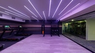 This single-floor fitness facility is designed with a contemporary aesthetic, combining high-performance equipment with striking architectural details. The main workout area is defined by its dramatic ceiling installation of linear LED strip lighting arranged in a dynamic, angular pattern that bathes the space in an atmospheric purple hue, creating a motivating and energetic environment.

The primary training zone features light wood-style flooring, dark accent walls with a horizontal striped texture, and a robust functional training rig equipped with storage for fitness accessories. Two commercial-grade treadmills with integrated touchscreens are positioned along the feature wall, accompanied by rubber flooring for impact absorption.

A glass-partitioned section houses additional cardio equipment, including stationary bikes with interactive displays, maintaining an open and visually connected feel throughout the facility. This area transitions into a secondary corridor space with light walls, recessed lighting, and built-in shelving, providing practical storage solutions.

The overall design balances bold aesthetics with functional layout, utilizing glass walls to maximize sightlines across zones. The cohesive use of dark tones, ambient LED lighting, and quality finishes throughout gives the facility a polished, boutique gym atmosphere suited for a focused fitness experience.