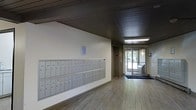 This single-floor space serves as a welcoming lobby and mail area, designed with practicality and clean aesthetics in mind. The layout is straightforward yet functional, anchored by a large bank of individual metal mailboxes mounted along one wall — providing convenient mail access for numerous occupants. The flooring consists of light wood-tone planks that run the length of the corridor, contributing to a bright and open feel. Overhead, a dark wood-paneled ceiling adds a layer of architectural contrast to the lighter walls and floor, with recessed and flush-mounted light fixtures ensuring the space is well illuminated. Toward the end of the hallway, a glass-panel entrance door allows natural light to filter in, offering a view of the greenery and exterior surroundings beyond. A door accessing an elevator is also visible, indicating connectivity to other levels of the building. A small window positioned above the mailboxes provides additional natural light. A dark entrance mat is placed near the main door, adding a practical touch. Overall, the space strikes a balance between utilitarian design and a welcoming atmosphere, serving efficiently as a transitional entry point.
