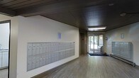 This single-floor space serves as a welcoming lobby and mail area, designed with practicality and clean aesthetics in mind. The layout is straightforward yet functional, anchored by a large bank of individual metal mailboxes mounted along one wall — providing convenient mail access for numerous occupants. The flooring consists of light wood-tone planks that run the length of the corridor, contributing to a bright and open feel. Overhead, a dark wood-paneled ceiling adds a layer of architectural contrast to the lighter walls and floor, with recessed and flush-mounted light fixtures ensuring the space is well illuminated. Toward the end of the hallway, a glass-panel entrance door allows natural light to filter in, offering a view of the greenery and exterior surroundings beyond. A door accessing an elevator is also visible, indicating connectivity to other levels of the building. A small window positioned above the mailboxes provides additional natural light. A dark entrance mat is placed near the main door, adding a practical touch. Overall, the space strikes a balance between utilitarian design and a welcoming atmosphere, serving efficiently as a transitional entry point.