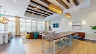 Located at 8300 Merrill Rd in Jacksonville, FL, this thoughtfully designed community office space blends modern functionality with warm, coastal-inspired aesthetics. The single-floor layout is unified by striking exposed dark wood ceiling beams that run the length of the space, paired with recessed lighting and oversized woven pendant fixtures that add texture and character overhead. Light hardwood flooring flows throughout the main gathering areas, grounding the bright, airy atmosphere created by large multi-pane windows that invite generous natural light and outdoor views. The lounge area is anchored by a teal sectional sofa, accent chairs in neutral tones, and layered area rugs, all arranged around a wall-mounted flat-screen TV — ideal for relaxed gatherings or informal meetings. A long communal high-top table with mesh chairs serves as a central workspace or dining area. The kitchen is a standout feature, appointed with crisp white shaker cabinetry, quartz countertops, a large island with bar seating, stainless steel appliances including a double wall oven and refrigerator, and an eye-catching mosaic tile accent wall. Decorative lantern-style pendant lights complete the kitchen's refined look. Throughout the space, carefully chosen furnishings and greenery create a welcoming, upscale ambiance well-suited for work and community connection.