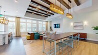 Located at 8300 Merrill Rd in Jacksonville, FL, this thoughtfully designed community office space blends modern functionality with warm, coastal-inspired aesthetics. The single-floor layout is unified by striking exposed dark wood ceiling beams that run the length of the space, paired with recessed lighting and oversized woven pendant fixtures that add texture and character overhead. Light hardwood flooring flows throughout the main gathering areas, grounding the bright, airy atmosphere created by large multi-pane windows that invite generous natural light and outdoor views. The lounge area is anchored by a teal sectional sofa, accent chairs in neutral tones, and layered area rugs, all arranged around a wall-mounted flat-screen TV — ideal for relaxed gatherings or informal meetings. A long communal high-top table with mesh chairs serves as a central workspace or dining area. The kitchen is a standout feature, appointed with crisp white shaker cabinetry, quartz countertops, a large island with bar seating, stainless steel appliances including a double wall oven and refrigerator, and an eye-catching mosaic tile accent wall. Decorative lantern-style pendant lights complete the kitchen's refined look. Throughout the space, carefully chosen furnishings and greenery create a welcoming, upscale ambiance well-suited for work and community connection.