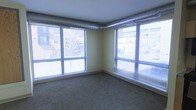 This single-floor space offers a clean, open layout with an industrial-modern character defined by its standout architectural details. Large corner windows on two walls flood the interior with natural light and frame views of the surrounding urban streetscape, including neighboring buildings and street-level activity. The dual-window configuration creates an airy, expansive feel and provides excellent daylight penetration throughout the day. Exposed metallic ductwork runs along the ceiling perimeter, lending the space an industrial aesthetic that complements its urban setting. The flooring consists of durable, neutral-toned tile laid throughout, offering both practicality and a clean visual foundation. Light yellow-toned walls contribute to a bright and open ambiance, enhancing the natural light brought in by the oversized windows. Horizontal blinds on the windows provide adjustable light control and privacy as needed. The space presents a versatile, straightforward layout well-suited for a variety of uses, with its combination of abundant natural light, urban views, and utilitarian finishes making it an adaptable and functional environment.