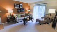 This single-floor model space showcases a thoughtfully designed living area that balances comfort and style. The living room is anchored by a bold terracotta-toned accent wall, which adds warmth and visual depth to the room. Soft, wall-to-wall carpeting extends throughout the space, contributing to a cozy and welcoming atmosphere.

The room accommodates generous seating, including a light-toned sofa dressed with decorative throw pillows in earthy greens and warm reds, complemented by an upholstered chair and ottoman. A dark wood octagonal coffee table serves as a functional centerpiece, styled with decorative accents that enhance the overall aesthetic.

A standout feature of the living area is the large sliding glass door fitted with vertical blinds, which allows abundant natural light to fill the room while offering a view of the outdoor area beyond. This connection to the exterior adds an airy, open quality to the space.

Wall-mounted artwork and a softly glowing table lamp further enhance the inviting ambiance, making the living area feel both curated and livable. The overall design reflects a warm, neutral palette with carefully chosen furnishings suited to everyday comfort.
