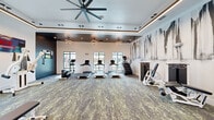 Located at 4535 SW 54th St Rd in Ocala, FL, this well-appointed fitness center offers a thoughtfully designed, single-floor layout packed with a comprehensive range of exercise equipment. The space features a striking tray ceiling accented with warm LED strip lighting and complemented by modern sculptural pendant lights and a large ceiling fan, creating an energizing yet refined atmosphere.

The cardio zone is lined with multiple treadmills, an elliptical, and a stair climber, all positioned in front of large, multi-pane windows that flood the space with natural light and offer views of the outdoor pool and landscaping. A dedicated strength and functional training area showcases a UFC-branded cable resistance machine, a full dumbbell rack, a weight bench, and various selectorized strength machines.

The flooring throughout features a durable, textured carpet tile in earthy tones, adding comfort underfoot while complementing the contemporary aesthetic. Decorative wall murals with bold, abstract brushstroke designs serve as artistic focal points, while motivational athletic photography lines the upper walls. Mirrored panels further enhance the sense of openness. The overall design balances high-end aesthetics with practical functionality, making this fitness center a premium amenity for the surrounding community.