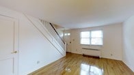 This two-floor space offers a clean, classic layout with notable architectural details. The main level entry area showcases gleaming hardwood floors that reflect natural light, contributing to a bright and open atmosphere. A white-painted staircase with a dark wood handrail and traditional balustrade rises prominently along one wall, serving as a functional yet visually striking feature of the main level.

The front-facing area includes a solid entry door with a transom sidelite window, allowing additional natural light into the entryway. A double-pane window on the adjacent wall further enhances brightness and provides views of the surrounding greenery outside. A cast-iron radiator sits beneath the window, indicating a hot-water heating system that provides consistent warmth throughout the colder months.

The walls are finished in crisp white throughout, creating a neutral and versatile backdrop. The polished hardwood flooring extends seamlessly across the main level, adding warmth and continuity. Overall, the space maintains a tidy, understated aesthetic with a traditional residential character, offering a solid foundation that is both functional and inviting.