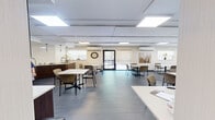 Located at 840 Trillium Park in Sarnia, Ontario, this single-floor communal space is thoughtfully designed to serve as both a dining and social gathering area. The open layout accommodates multiple dining tables with comfortable upholstered chairs arranged throughout, creating a welcoming and functional environment for group use.

The space is finished with large-format grey tile flooring and a neutral warm-beige color palette on the walls, contributing to a calm and inviting ambiance. Flush panel ceiling lights provide consistent, bright illumination throughout, complemented by natural light streaming in through several windows. A glass door at one end opens directly to an outdoor patio area, enhancing the connection to the exterior greenspace.

A fully equipped kitchen anchors one end of the room, featuring dark espresso cabinetry, a central island, pendant lighting, and ample counter space — well-suited for meal preparation and service. A wall-mounted flat-screen television set within a dark wood surround serves as a focal point in the lounge area, adding a social and entertainment element to the space. Decorative artwork is displayed along the walls, adding warmth and character. A split-unit air conditioner ensures year-round comfort. The space also includes a hallway with a clearly marked exit, supporting safe and efficient circulation.