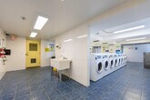 Located at 1577 Lawrence Ave W in Toronto's M6L neighbourhood, this communal laundry facility is a single-floor space offering a practical and well-organized environment for residents. The layout is efficiently designed, with multiple rows of front-loading washing machines and dryers arranged along the walls, providing ample capacity for high-volume use. The interior features white large-format wall tiles that lend a clean, hygienic aesthetic throughout, complemented by blue speckled floor tiles that add a subtle contrast to the bright surroundings. Overhead, recessed fluorescent lighting panels illuminate the space evenly, ensuring clear visibility at all times. A utility sink with a freestanding basin is conveniently positioned for pre-treatment or general use, while a long laminate folding counter offers residents a dedicated surface for sorting and folding laundry. A bulletin board mounted on one wall serves as an informational hub, displaying notices and instructions for users. Seating is available near the entrance, providing a comfortable waiting area. Safety features include a fire extinguisher and security camera. Access is through a windowed yellow door, and the overall space maintains a tidy, functional character suited to shared residential use.