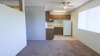 This single-floor space offers a practical and functional layout centered around an open-concept living and kitchen area. The kitchen is equipped with warm-toned wood upper and lower cabinetry, a countertop workspace, a built-in sink, a stovetop range, and a full-size white refrigerator, providing ample storage and utility for everyday use. Light tile flooring in the kitchen transitions seamlessly into plush carpeting in the adjacent living area, creating a clear yet open distinction between the two spaces.

A ceiling fan with an integrated light fixture is centrally positioned overhead, ensuring air circulation and ambient lighting throughout. A fluorescent under-cabinet light above the kitchen workspace adds task lighting for food preparation. The walls are finished in a neutral, muted tone that complements the overall understated aesthetic of the space, while a textured ceiling adds a subtle architectural detail. The layout is efficient and straightforward, making good use of the available square footage. Overall, the space presents a clean, no-frills environment well-suited for comfortable everyday living.
