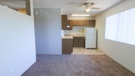 This single-floor space offers a practical and functional layout centered around an open-concept living and kitchen area. The kitchen is equipped with warm-toned wood upper and lower cabinetry, a countertop workspace, a built-in sink, a stovetop range, and a full-size white refrigerator, providing ample storage and utility for everyday use. Light tile flooring in the kitchen transitions seamlessly into plush carpeting in the adjacent living area, creating a clear yet open distinction between the two spaces.

A ceiling fan with an integrated light fixture is centrally positioned overhead, ensuring air circulation and ambient lighting throughout. A fluorescent under-cabinet light above the kitchen workspace adds task lighting for food preparation. The walls are finished in a neutral, muted tone that complements the overall understated aesthetic of the space, while a textured ceiling adds a subtle architectural detail. The layout is efficient and straightforward, making good use of the available square footage. Overall, the space presents a clean, no-frills environment well-suited for comfortable everyday living.
