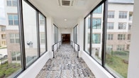 Located on Henry Parkway in Flower Mound, Texas, this single-level connector corridor serves as a distinctive transitional passageway between buildings within a larger multi-story complex. The corridor's most striking design element is its elaborately patterned tile flooring, featuring an intricate blue, grey, and white motif that runs the full length of the passage, lending it a refined, decorative character uncommon in utility spaces.

Floor-to-ceiling black-framed windows line both sides of the corridor continuously, flooding the interior with abundant natural light and offering clear sightlines to the surrounding building exteriors and landscaped outdoor areas. The white-painted walls and clean-lined ceiling — equipped with recessed lighting and ceiling-mounted HVAC units — create a bright, well-maintained environment throughout.

Accessibility is thoughtfully integrated into the design, with black steel handrails flanking a centrally positioned ramp that gently slopes along the corridor's length, ensuring ease of movement for all users. Exit signage is clearly positioned at the far end of the passage, which opens into an adjoining interior space. The overall ambiance is modern and polished, balancing functional building connectivity with intentional architectural detail and an abundance of natural light.