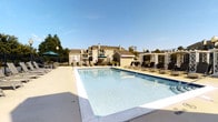 This outdoor amenity area centers around a generously sized rectangular swimming pool with clear, blue water and a wide surrounding deck. The pool deck is finished in light-colored concrete, providing ample space for lounging and circulation. Rows of dark-framed chaise lounge chairs are arranged along the poolside, offering residents dedicated sunbathing areas. Several umbrella-shaded seating stations are positioned around the perimeter, providing relief from direct sunlight. A decorative white pergola anchors one end of the deck, adding a classic architectural accent to the outdoor space. Wrought iron-style fencing encloses the pool area, defining its boundary while maintaining open sightlines to the surrounding landscape. Mature trees and greenery frame the space, contributing to a pleasant, park-like atmosphere. The backdrop features a multi-story residential building with balconies and neutral-toned siding, visible under an open, clear blue sky. The overall ambiance is bright, clean, and well-maintained, making the pool area a functional and inviting outdoor retreat suited for warm-weather relaxation and recreation.