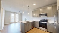 This efficiently designed one-bedroom, one-bathroom residence offers a thoughtful single-floor layout that maximizes both functionality and comfort. The open-concept kitchen and living area serves as the heart of the home, featuring warm light hardwood flooring throughout. The kitchen is outfitted with sleek gray shaker-style cabinetry, a light-toned countertop peninsula with an undermount sink, and a full suite of stainless steel appliances including a refrigerator, electric range, built-in microwave, and dishwasher. Dual pendant lights hang above the peninsula, while a ceiling fan overhead serves the adjacent living area, which receives natural light through large windows with blinds. The entry hallway provides a clean transition from the front door into the main living space. The bedroom offers a comfortable, carpeted retreat with a ceiling fan, a bright window with natural light, and neutral wall tones that create a calm atmosphere. Two closets provide ample storage. The bathroom features wood-look flooring, a tub-shower combination with tiled surround, and direct access to a well-organized closet. Throughout, the space maintains a clean, modern aesthetic with a neutral color palette and consistent finishes.