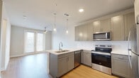 This efficiently designed one-bedroom, one-bathroom residence offers a thoughtful single-floor layout that maximizes both functionality and comfort. The open-concept kitchen and living area serves as the heart of the home, featuring warm light hardwood flooring throughout. The kitchen is outfitted with sleek gray shaker-style cabinetry, a light-toned countertop peninsula with an undermount sink, and a full suite of stainless steel appliances including a refrigerator, electric range, built-in microwave, and dishwasher. Dual pendant lights hang above the peninsula, while a ceiling fan overhead serves the adjacent living area, which receives natural light through large windows with blinds. The entry hallway provides a clean transition from the front door into the main living space. The bedroom offers a comfortable, carpeted retreat with a ceiling fan, a bright window with natural light, and neutral wall tones that create a calm atmosphere. Two closets provide ample storage. The bathroom features wood-look flooring, a tub-shower combination with tiled surround, and direct access to a well-organized closet. Throughout, the space maintains a clean, modern aesthetic with a neutral color palette and consistent finishes.