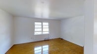 This single-floor space presents a clean, minimalist aesthetic that is both functional and inviting. The interior is characterized by smooth, white-painted walls and ceilings that create a bright, airy atmosphere throughout. Polished hardwood floors in a warm honey tone run throughout the space, reflecting natural light and adding a timeless charm to the overall design. A double-pane window serves as a key architectural feature, allowing generous amounts of natural light to fill the room while offering views of the surrounding greenery outdoors. The white walls and ceiling work in harmony with the light hardwood flooring to maximize the sense of openness and space. The layout is straightforward and adaptable, making it well-suited for a variety of uses and furniture arrangements. Baseboard details run along the perimeter of the room, adding a subtle finishing touch to the otherwise understated interior. The space maintains a neutral palette throughout, providing an excellent canvas for personalization. Overall, this space combines practical design with a light-filled, welcoming ambiance that emphasizes simplicity and versatility.