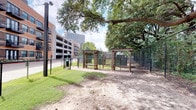 This small dog park is a purpose-built outdoor amenity designed to provide a safe and engaging environment for small dogs. The park is fully enclosed by a sturdy black chain-link fence, ensuring pets remain secure during off-leash activity. A charming wood pergola marks the entrance, flanked by lattice panels that add architectural character to the space. A brick paver pathway leads visitors from the entry point through the park, offering a clean and defined walkway. The grounds feature a mix of open dirt play areas and patches of grass, providing varied terrain for exploration. Mature oak trees with broad, spreading canopies dominate the landscape, delivering generous natural shade throughout the park. The lush tree coverage creates a cool, sheltered atmosphere even on sunny days. The park is situated within an urban residential setting, with multi-story buildings visible along the perimeter, placing it conveniently close to the surrounding community. A waste station is available near the entrance, supporting responsible pet ownership. The overall ambiance is green and natural, offering a pleasant retreat for small dogs and their owners within an active urban neighborhood.
