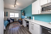 Located at 4117 University Ave in Cedar Falls, Iowa, this efficiently designed studio unit makes smart use of its compact footprint across a single floor. The open-plan layout seamlessly combines the kitchen and living area, creating a cohesive and functional living space. A vivid teal accent wall serves as the visual centerpiece, lending the unit a fresh and modern character that is complemented by crisp white cabinetry and light wood-tone flooring throughout.

The kitchen area features white upper and lower cabinets, a smooth countertop workspace, and built-in appliances including a microwave, offering practical everyday convenience. The living area is brightened by a large window that admits generous natural light and provides outward views, while a wall-mounted flat-screen TV and track lighting overhead add to the unit's functional appeal. A colorful area rug and seating area complete the inviting atmosphere.

A cleverly integrated Murphy bed folds down from a white wall panel, maximizing floor space when not in use and keeping the layout versatile. The teal entry door adds a bold design touch upon arrival.

The bathroom is cleanly appointed with white cabinetry, a single vanity with sink, and a walk-in shower stall, accented with teal details that tie into the overall color palette. The textured ceiling and wood-look flooring carry through the space consistently, reinforcing a cohesive, well-considered design.