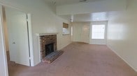 This single-floor space offers an open and functional layout with a warm, understated character. The living area serves as the centerpiece of the home, anchored by a traditional brick fireplace with a white-painted mantel, adding a classic architectural detail that draws the eye and gives the room a cozy focal point. Neutral-toned carpet flows throughout, providing a soft and cohesive foundation across the open floor plan.

The ceiling features a recessed section toward the front of the space, adding subtle visual interest and a sense of dimension to the layout. A large window near the front entry allows natural light to fill the room, brightening the neutral cream-colored walls. The entryway is framed by a paneled white door, contributing to a clean, traditional aesthetic.

Toward the rear of the living space, a pass-through window connects to what appears to be a kitchen area, offering convenient sightlines and promoting an open feel between spaces. Recessed lighting in the ceiling provides consistent illumination throughout. Overall, the space presents a straightforward, functional design well-suited for comfortable everyday living.
