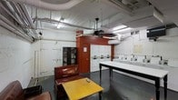 This single-floor communal laundry space combines practical utility with a modest lounge area, making it a functional hub for residents. The room features exposed pipes and ductwork along the ceiling, lending an industrial character to the space. Overhead fluorescent lighting provides ample illumination throughout, supplemented by a ceiling fan that aids air circulation. The walls are painted in a contrast of white brick and a bold terracotta-red accent, adding visual interest to the utilitarian environment.

Along one wall, a row of stacked washing machines and dryers offers multiple laundry stations, each equipped with individual controls for ease of use. A large folding table positioned centrally provides generous workspace for sorting and folding laundry. A dark bookcase near the entrance offers additional storage and a small reading selection.

The lounge area, set apart from the laundry stations, includes a cushioned armchair and a wooden coffee table, offering a comfortable waiting space. The concrete flooring is practical and easy to maintain, consistent with the room's overall utilitarian design. The space is efficiently organized to balance laundry functionality with basic comfort amenities.