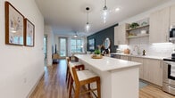 Located at 9821 Research Drive in Charlotte, NC, this thoughtfully designed one-bedroom, two-bathroom unit offers a modern and cohesive living experience across a single floor. The open-concept layout seamlessly connects the kitchen and living room, creating an inviting, functional space. The kitchen is outfitted with light wood cabinetry, white subway tile backsplash, stainless steel appliances — including a range, microwave, dishwasher, and refrigerator — and a white quartz island with bar seating, illuminated by stylish pendant lights. The living room features warm hardwood-style flooring, a bold teal accent wall, a ceiling fan, and large windows with a glass balcony door that fill the space with natural light and offer outdoor views. The bedroom is a comfortable retreat highlighted by a striking decorative wallpapered accent wall, a large window, and direct access to a well-appointed en-suite bathroom. The primary bathroom includes a light wood vanity, quartz countertop, soaking tub, and tiled shower. A second full bathroom and a dedicated laundry room with a stacked washer/dryer unit and wire shelving add practicality. Throughout, the design maintains a clean, contemporary aesthetic with neutral tones and quality finishes.