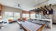Located at 1818 Park Ave in Dallas, TX, this generously sized amenity lounge spans a single floor and delivers a sophisticated, urban aesthetic throughout. The main gathering area makes an immediate impression with its exposed brick accent walls, dark wood beam ceiling details, and an array of large windows that flood the space with natural light while framing sweeping views of the Dallas skyline. A full-size pool table anchors the center of the room, complemented by comfortable sectional seating and swivel lounge chairs arranged for social gatherings. A stylish wet bar with mosaic tile backsplash, pendant lighting, and bar seating serves as a focal point, while stainless steel appliances and a built-in kitchen area add functional convenience. High-top tables and bar stools near the windows provide additional seating with city views. A separate private meeting or conference room features a striking full-wall photographic mural, a round table with upholstered chairs, and a dramatic starburst chandelier overhead. The entry corridor is accented with bold blue walls, curated artwork, and elevator access. A rooftop terrace with wood-look tile flooring, synthetic turf, and sleek metal railings offers unobstructed panoramic views of the Dallas downtown skyline, completing this well-appointed communal space.