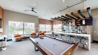 Located at 1818 Park Ave in Dallas, TX, this generously sized amenity lounge spans a single floor and delivers a sophisticated, urban aesthetic throughout. The main gathering area makes an immediate impression with its exposed brick accent walls, dark wood beam ceiling details, and an array of large windows that flood the space with natural light while framing sweeping views of the Dallas skyline. A full-size pool table anchors the center of the room, complemented by comfortable sectional seating and swivel lounge chairs arranged for social gatherings. A stylish wet bar with mosaic tile backsplash, pendant lighting, and bar seating serves as a focal point, while stainless steel appliances and a built-in kitchen area add functional convenience. High-top tables and bar stools near the windows provide additional seating with city views. A separate private meeting or conference room features a striking full-wall photographic mural, a round table with upholstered chairs, and a dramatic starburst chandelier overhead. The entry corridor is accented with bold blue walls, curated artwork, and elevator access. A rooftop terrace with wood-look tile flooring, synthetic turf, and sleek metal railings offers unobstructed panoramic views of the Dallas downtown skyline, completing this well-appointed communal space.