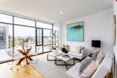 This single-floor residence is defined by a clean, contemporary design with an open layout that connects the kitchen, dining, and living areas into one cohesive space. Warm hardwood flooring flows throughout, complementing the bright white walls and recessed lighting overhead.

The kitchen is outfitted with flat-front white cabinetry extending to the ceiling, a marble-style backsplash, light stone countertops, a gas cooktop, double wall ovens, and an integrated refrigerator. A glass-top dining table with wire-frame chairs occupies the adjacent dining area, positioned beside floor-to-ceiling windows that frame sweeping city views.

The living room benefits greatly from these large-format windows, which flood the space with natural light and provide an elevated urban panorama. The room accommodates generous seating and features notable ceiling height, enhancing the overall sense of openness.

The bedroom is a calm, softly lit retreat with smooth white walls and hardwood flooring. The bathroom is finished with large-format neutral tile covering the walls and floors, a soaking tub with shower, a wall-mounted white vanity, and a backlit mirror, delivering a polished, spa-like aesthetic throughout.
