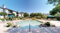 This expansive outdoor pool area offers a resort-inspired retreat designed for relaxation and recreation. The centerpiece is a generously sized swimming pool with clear, blue-green water, framed by a well-maintained aggregate and paver sundeck that provides ample space for lounging. Multiple reclining chairs are arranged poolside, accompanied by teal market umbrellas that offer shaded seating throughout the deck. Colorful flowering plants and neatly trimmed hedges line the pool's perimeter, adding vibrant seasonal color and polished landscaping detail. Dedicated seating areas feature round glass-top tables with umbrella coverage, providing comfortable spots for outdoor dining or casual gatherings. Mature trees throughout the surrounding grounds contribute significant natural shade and a lush, park-like ambiance. A charming clubhouse building anchors one end of the pool, featuring a pitched roof, classic architectural detailing, and direct access to the pool deck. The entire area is enclosed by a wrought iron fence, ensuring privacy and security. Elevated residential buildings visible in the background provide a sense of community scale. Under clear blue skies, this outdoor space delivers a bright, welcoming atmosphere ideal for year-round outdoor enjoyment.